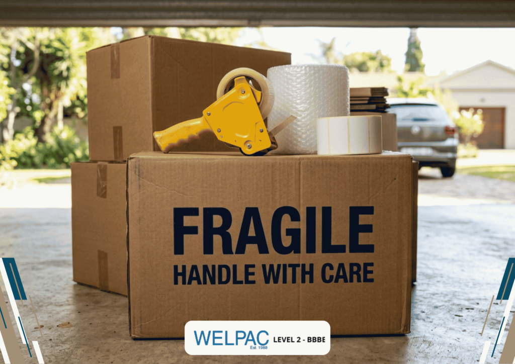 Three cardboard boxes, one labeled “FRAGILE HANDLE WITH CARE,” with packing tape, bubble wrap, and labels on top. A car and houses are visible in the blurred background.